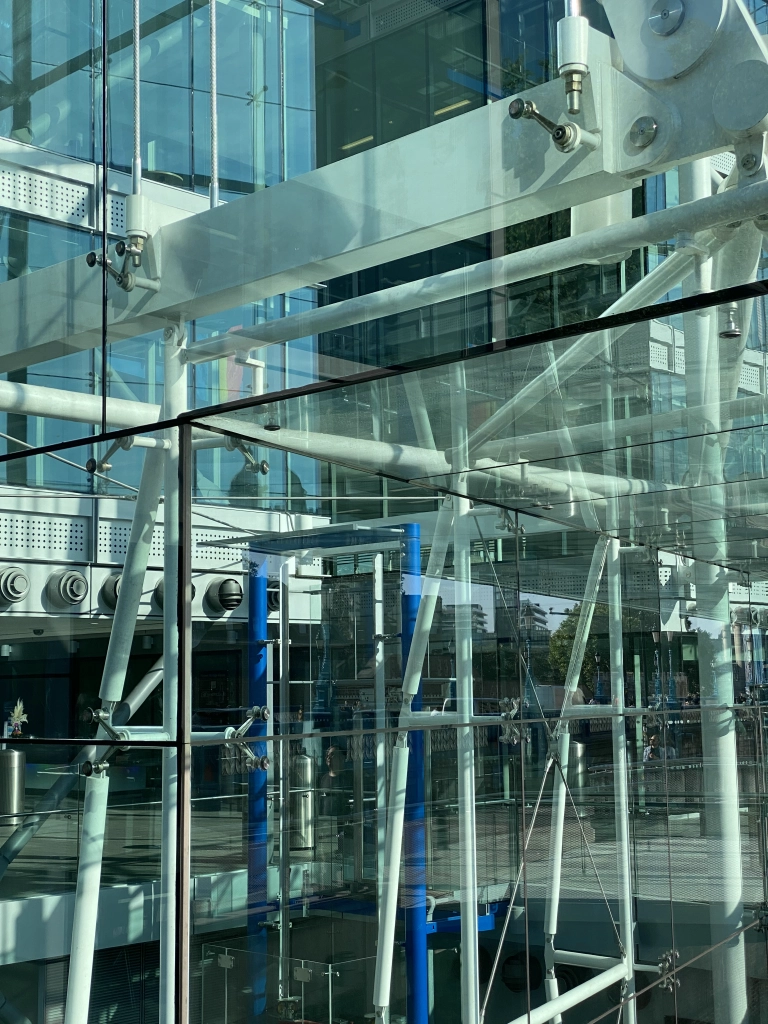 An external all-glass elevator and accompanying metal supports create a chaotic mess of reflections with themselves and the outside street. A pride flag can be seen thru a gap.