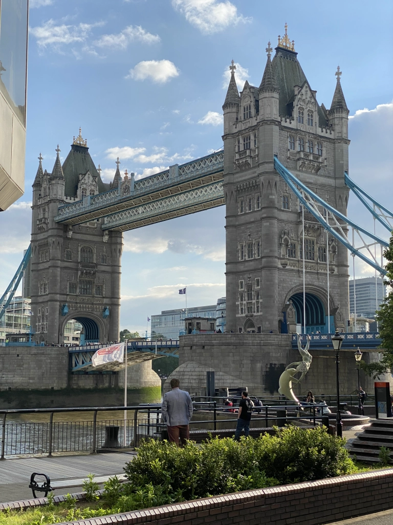 Tower Bridge in London as viewed from the north shore, with the Girl And Dolphin sculpture visible. A few pedestrians mill about the waterfront.